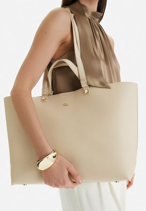 Donna con blusa marrone senza maniche e pantaloni bianchi che tiene una grande borsa a mano beige con texture, dettagli dorati e un braccialetto dorato massiccio al polso.