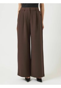 Donna che indossa pantaloni marroni a vita alta e gamba larga con tasche laterali e décolleté neri a punta, in piedi davanti a uno sfondo uniforme.