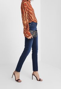 Blusa a rayas en tonos óxido con botones, jeans ajustados oscuros y bolso de mano negro acolchado. Modelo luciendo sandalias de tacón alto abiertas de color púrpura.