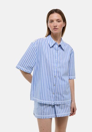 Femme à la peau claire et aux cheveux bruns lisses portant une chemise boutonnée à rayures verticales bleues et blanches et un short assorti sur fond uni.