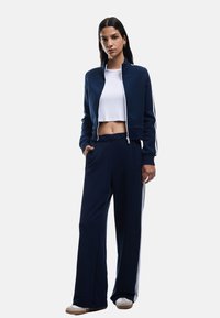 Chándal atlético azul marino que incluye una chaqueta ajustada con cremallera y pantalones de pierna ancha con rayas laterales, fabricado en una tela suave con una textura lisa.