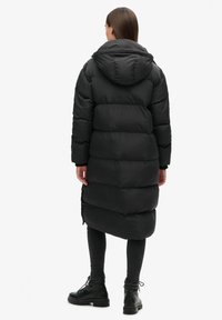 Zwarte pufferjas met een capuchon, voorzien van een dikke gewatteerde uitstraling en een knielange snit. G gedragen met donkere leggings en zwarte veterschoenen.