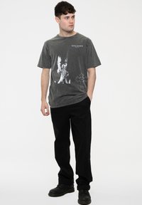Gray cotton t-shirt featuring a front graphic of a person with hands clasped, alongside text “TUPAC SHAKUR” and a cross design. Paired with black trousers.