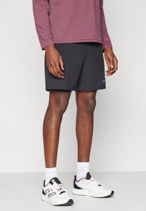 Person trägt schwarze Sportshorts, weiße New Balance Turnschuhe, weiße Socken und ein langärmliges lila Hemd und steht vor einem einfarbigen Hintergrund.