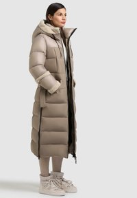 Cappotto puffer beige con un taglio lungo, colletto peloso e design trapuntato accentuato. Indossato con leggings e calzature coordinate beige.