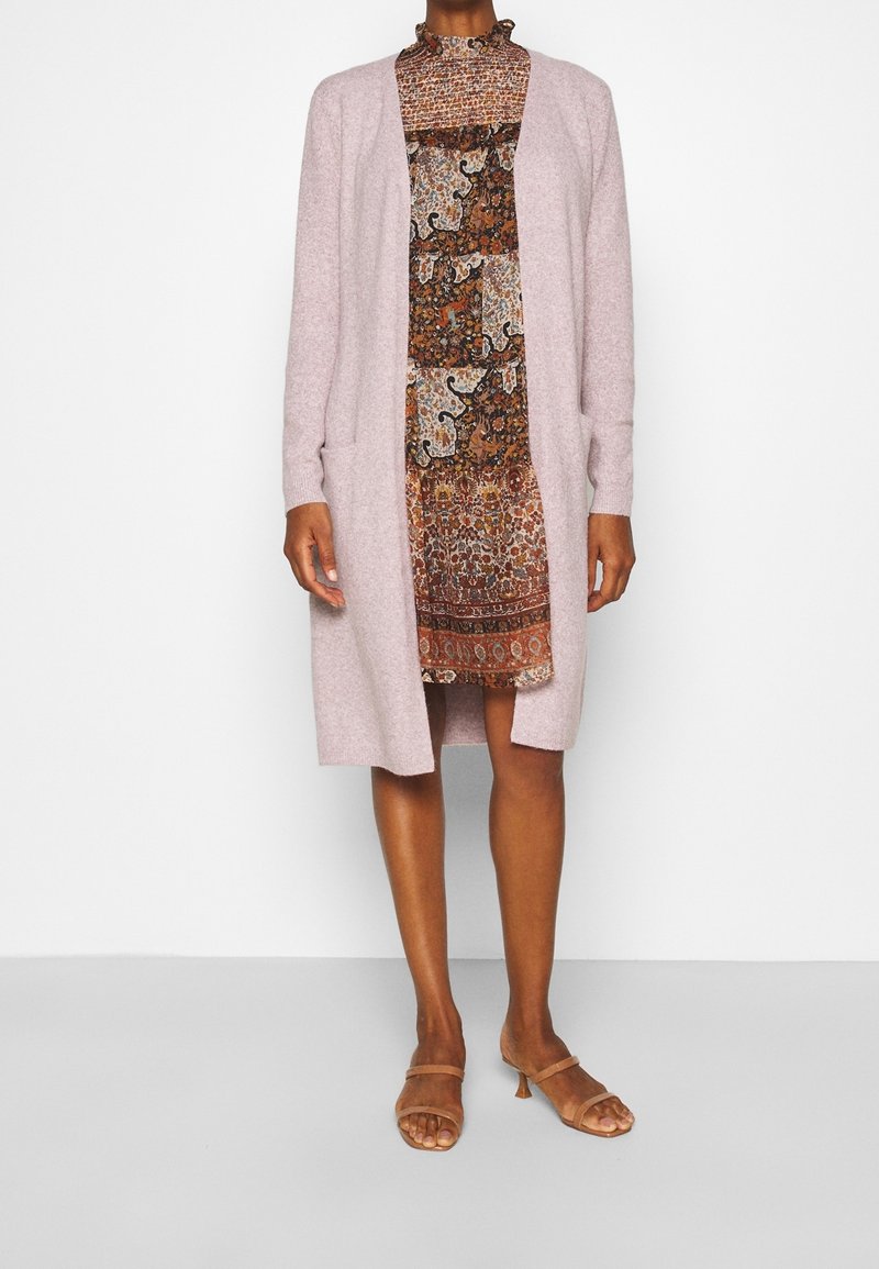 Femme portant un long cardigan rose clair sur une robe marron à motifs longueur genoux et des sandales à talons couleur camel, debout devant un fond blanc uni.
