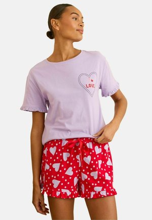 Nainen, joka käyttää laventelinväristä lyhythihaista toppia, jossa on sydän ja teksti "LOVE", yhdistettynä punaiseen shortseihin, joissa on valkoisia sydämiä ja pilkkuja, seisoo käsi lantiolla.