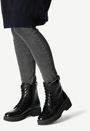Black lace-up boots made of patent leather, with a chunky sole and detailed stitching. Worn with grey skinny jeans.