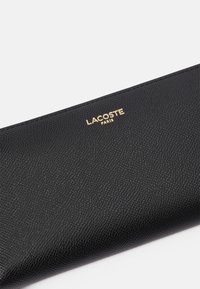 Portafoglio in pelle nera texturizzata con logo dorato "LACOSTE PARIS". Finitura liscia, forma rettangolare con un design elegante e senza hardware visibile.