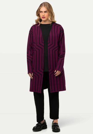 Langer lila Cardigan mit schwarzen geometrischen Streifen, Offene Vorderseite und langen Ärmeln. Getragen über einem schwarzen Oberteil und kombiniert mit schwarzen Hosen und Schuhen.