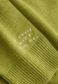 Tissu vert tricoté avec une texture côtelée, présentant un texte brodé en blanc : "Un petit oiseau m'a dit..." sur une section pliée.