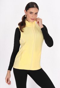 Vesta din fleece galben cu guler înalt și fermoar, purtată cu mâneci lungi negre și colanți. Dispune de buzunare laterale și o textură netedă.