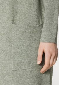 Grön stickad klänning med en texturerad yta, långa ärmar och en stor framficka på vänster sida. Tyget verkar mjukt och mysigt.