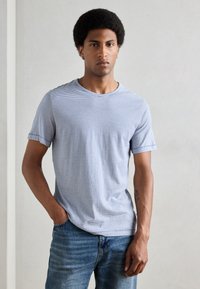 Tricou scurt cu maneci, în dungi gri și alb, realizat dintr-un material moale, asortat cu blugi din denim bleumarin. Modelul stă cu mâinile în buzunare.