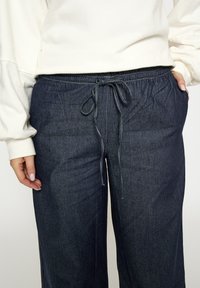 Tmavomodré džínové tepláky s elastickým pásom, nastaviteľným šnúrkou a bočnými vreckami. Hladká textúra a pohodlný strih.
