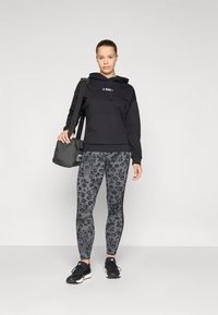 Felpa nera con cappuccio e logo bianco, abbinata a leggings grigi con stampa leopardata. Il modello indossa scarpe da ginnastica nere e tiene in mano una piccola borsa nera.