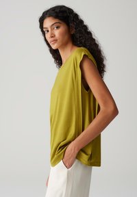 Top senza maniche di color verde oliva realizzato in un tessuto morbido, caratterizzato da spacco laterale e una vestibilità rilassata. Decorato con sottili dettagli drappeggiati.