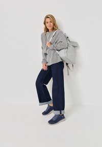 Sivi oversized pulover, tamne široke traperice s preokrenutim rubovima, sivi ruksak, mornarsko plavi tenisice s uzorkom, stojeći naslonjen uz bijeli zid.