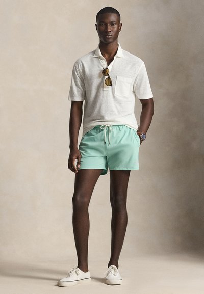 Νέος άνδρας στέκεται, φοράει λευκό πόλο πουκάμισο, σορτς μέντας πράσινου χρώματος με κορδόνι, λευκά αθλητικά παπούτσια, ρολόι και γυαλιά ηλίου κρεμασμένα από το πουκάμισο.