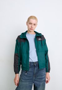 Teal en zwarte windbreaker met capuchon, elastische manchetten en een ritssluiting aan de voorkant. Bevat een logo op de borst en heeft een soepele textuur. G gedragen over een lichtblauw top.