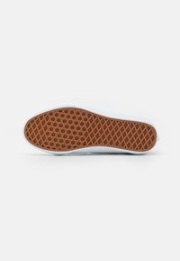 Shoe sole with brown waffle-pattern tread lies flat against a white surface with a white edge around the sole.