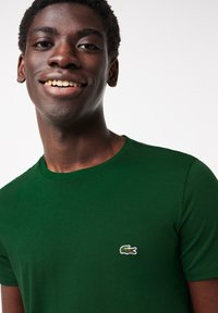 Vihreä puuvillapaita, jossa on pyöreä kaula-aukko ja lyhyet hihat. Paidassa on vasemmassa rinnassa pieni brodeerattu krokotiililogo.