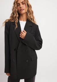 Musta kaksirivinen pikkutakki, jossa on leveät kaulukset, kaksi nappia, sivutaskut ja jämäkkä istuvuus, tyylitelty valkoisen paidan päällä.