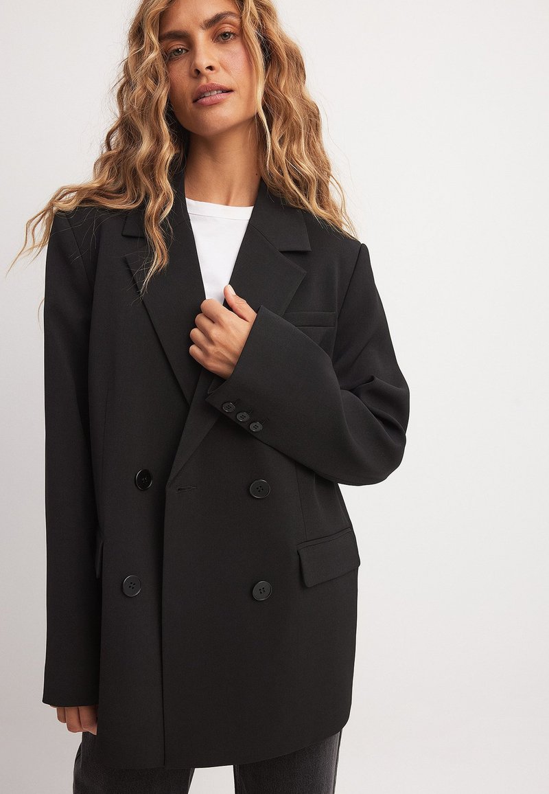 Musta kaksirivinen pikkutakki, jossa on leveät kaulukset, kaksi nappia, sivutaskut ja jämäkkä istuvuus, tyylitelty valkoisen paidan päällä.