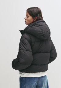 Ženska z valovitimi temnimi lasmi, oblečena v črno puhovko z ruto in modre kavbojke, stoji bočno ob preprostem ozadju.