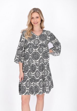 Uśmiechnięta kobieta o blond włosach, ubrana w czarno-białą sukienkę w kwiatowy wzór, sięgającą kolan z rękawami do połowy przedramienia, stojąca na białym tle.