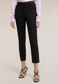 Vrouw draagt zwarte, op maat gemaakte enkellange broek, een lichtpaarse blouse en zwarte sandalen met hoge hak en gekruiste bandjes.