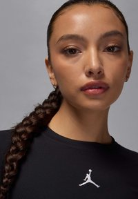 Sweat-shirt noir avec un col rond et un logo Jordan blanc, porté par une personne avec une coiffure tressée et des boucles d'oreilles créoles.