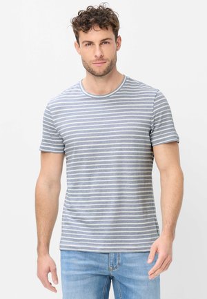 Mand med krøllet hår iført en kortærmet, lysegrå og hvid t-shirt med horisontale striber og lyseblå jeans, står mod en hvid baggrund.