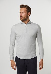 Graues langärmliges Poloshirt mit Kragen, drei vorderen Knöpfen und glatter Textur. Kombiniert mit dunklen Hosen steht das Modell vor einem neutralen Hintergrund.