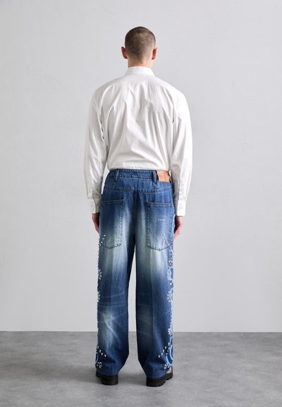 Hellblaue Jeans mit Verzierungen und Taschen, kombiniert mit einem weißen Langarmshirt. Gerade Passform. Schwarze Schuhe.