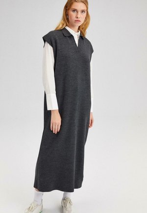 Abito grigio senza maniche con colletto, vestibilità comoda, realizzato in tessuto morbido, arriva alla caviglia, indossato sopra una camicia bianca e abbinato a sneaker.