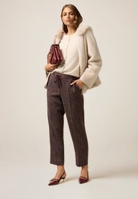 Beige faux fur-gekraagde jas, romige knitwear met knopen, bordeauxkleurige getextureerde broek met trekkoord en kastanjebruine lakleren hakken.