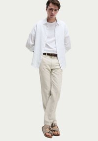 Camisa de lino blanca sobre una camiseta blanca, combinada con pantalones beige claros y sandalias grises con hebillas. Limpio, casual y con materiales texturizados.