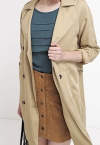 Manteau beige avec des boutons noirs sur un top plissé teal et une jupe en daim marron avec des boutons sur le devant. Un sac à main noir est tenu.