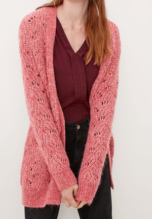 Femme aux longs cheveux roux portant un cardigan en maille rose texturé sur un chemisier bordeaux et un pantalon noir, debout, les mains jointes devant elle.