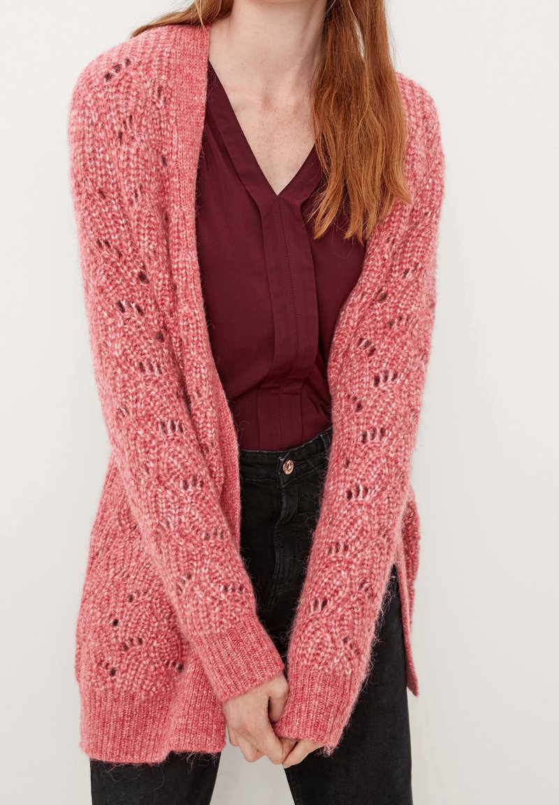 Femme aux longs cheveux roux portant un cardigan en maille rose texturé sur un chemisier bordeaux et un pantalon noir, debout, les mains jointes devant elle.