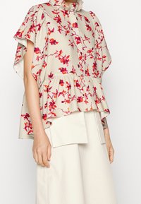 Donna che indossa una blusa floreale ampia con fiori rossi e rosa, orlo a volant, e pantaloni beige a gamba larga con grandi tasche frontali.