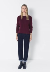 Femme blonde debout face à l’avant, portant un pull bordeaux à manches longues, un jean foncé retroussé et des bottines noires sur fond blanc.