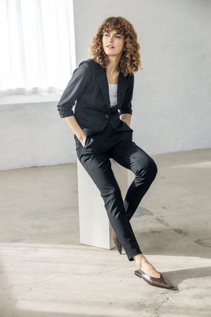 Kvinde med krøllet hår i sort jakkesæt og brune spidse flade sko, der sidder på en hvid blok i et minimalistisk rum med betongulv og væg.