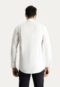 Camicia bianca a maniche lunghe con colletto alla francese, orlo curvo e polsini con bottone, realizzata in tessuto liscio, mostrata di spalle.