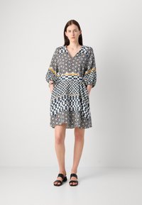 Kuvioitu mekko tummansinisessä, valkoisessa ja kultaisessa, jossa on V-pääntie, puffihihat ja taskut, sekä ilmava siluetti ja geometriset kuvioinnit.