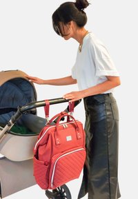 Bolsa de pañales acolchada roja con dos asas superiores y correas ajustables, sujeta a un carrito. Falda de cuero negra brillante, camiseta blanca de manga corta.