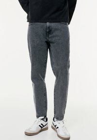 Jeans en denim gris avec une coupe décontractée, présentant une texture usée et des ourlets retroussés, associés à des chaussures de sport blanches et noires.