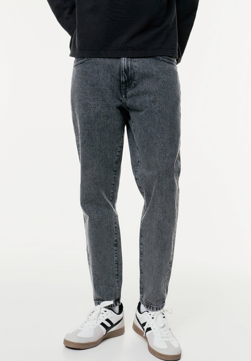 Jeans en denim gris avec une coupe décontractée, présentant une texture usée et des ourlets retroussés, associés à des chaussures de sport blanches et noires.