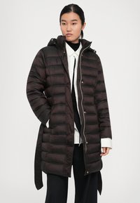 Ung kvinne iført en lang svart quilted dunjakke med hette over en hvit skjorte og svarte bukser, står med hendene i lommene.
